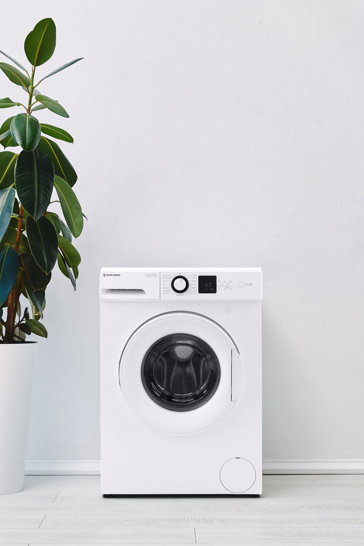 White washing machine next to a potted plant on a light wooden floor with a white wall background